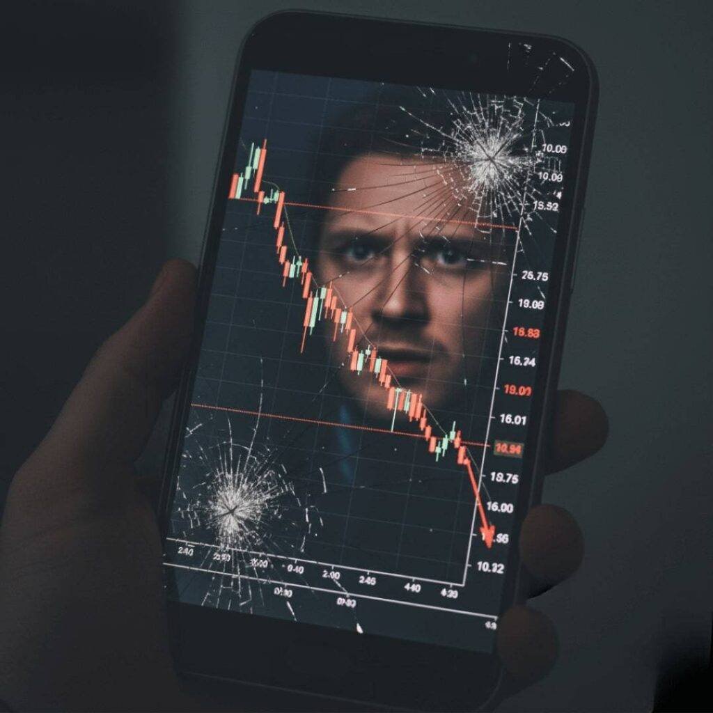 A cracked phone screen shows a red stock market dip with a stressed face reflected.